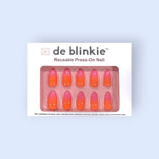 Product box shot of de blinkie Tropic Muse coral orange to hot pink ombre reusable press-on nails in medium almond displayed in white branded packaging on a pink tray against a soft blue background