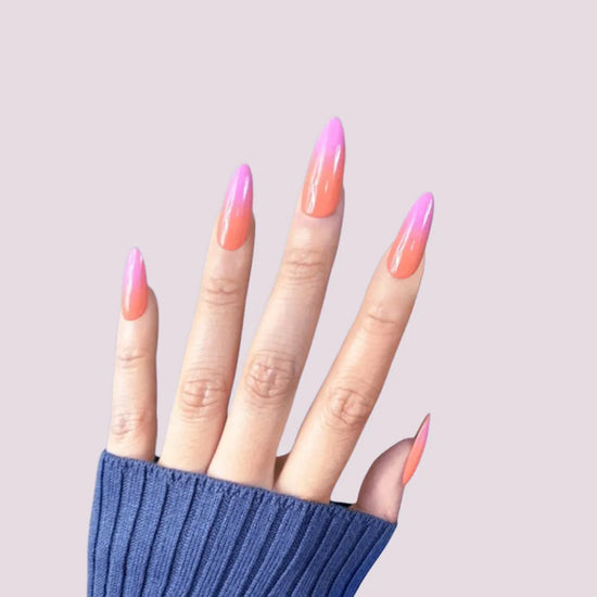 Spread hand showing de blinkie Tropic Muse coral orange to hot pink ombre press-on nails in medium almond against a soft lavender background with a blue ribbed knit sleeve