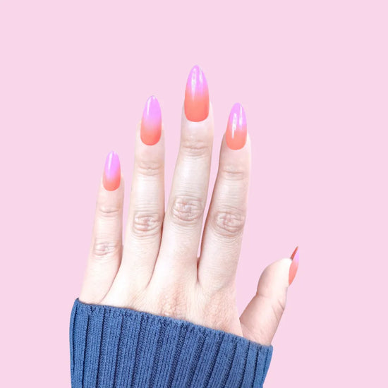 Spread hand showing de blinkie Tropic Muse coral orange to hot pink ombre press-on nails in medium almond against a bright pink background with a blue ribbed knit sleeve