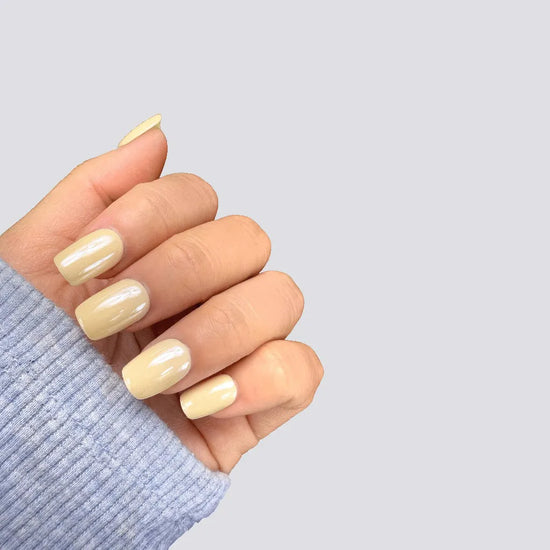 Claw pose showing de blinkie Vanilla Crème creamy butter yellow soft gloss short square press-on nails against light grey background with blue sleeve