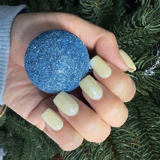 Hand holding blue glitter Christmas ornament showing de blinkie Vanilla Crème butter yellow cream short square press-on nails against festive pine tree background