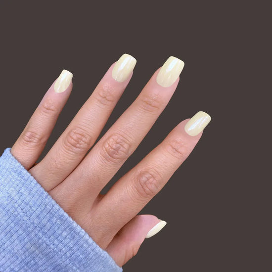 Spread hand showing de blinkie Vanilla Crème butter yellow cream short square press-on nails against dark brown background with light blue sleeve