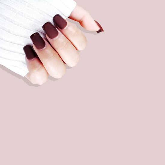 Claw pose showing de blinkie Velvet Bordeaux dark chocolate cherry velvet matte press-on nails in medium square shape against pink background with white ribbed knit sleeve