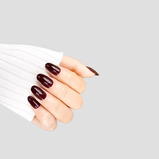 Diagonal claw pose showing de blinkie Velvet Noir dark bordeaux gloss press-on nails in short oval shape against grey background with white ribbed knit sleeve