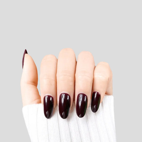Claw pose showing de blinkie Velvet Noir dark bordeaux gloss press-on nails in short oval shape against grey background with white ribbed knit sleeve