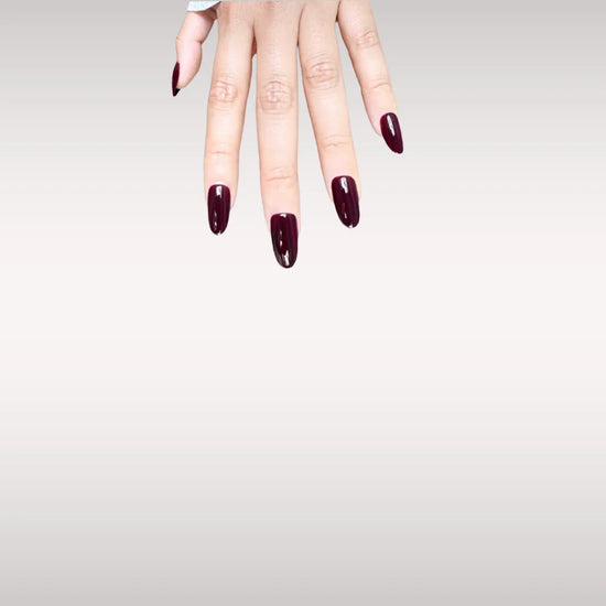 Fingers down pose showing de blinkie Velvet Noir dark bordeaux gloss press-on nails in short oval shape against off-white background with white ribbed knit sleeve