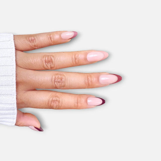 Horizontal spread hand showing de blinkie Velvet Vixen sheer blush base with alternating caramel, chocolate and black French tip press-on nails in short almond against a soft light grey background with a white ribbed knit sleeve