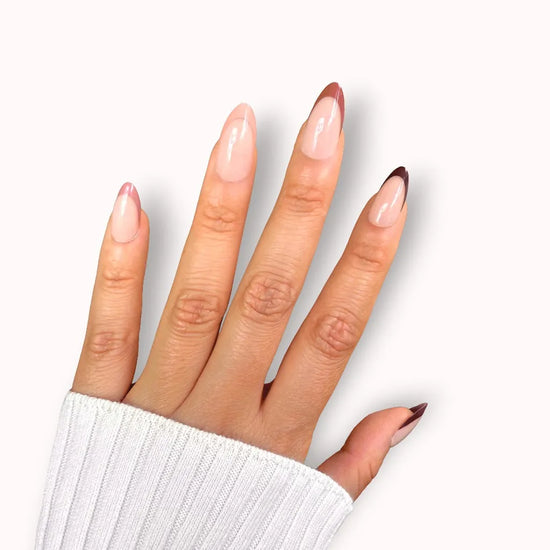 Spread hand showing de blinkie Velvet Vixen sheer blush base with alternating caramel, chocolate and black French tip press-on nails in short almond against a clean white background with a white ribbed knit sleeve